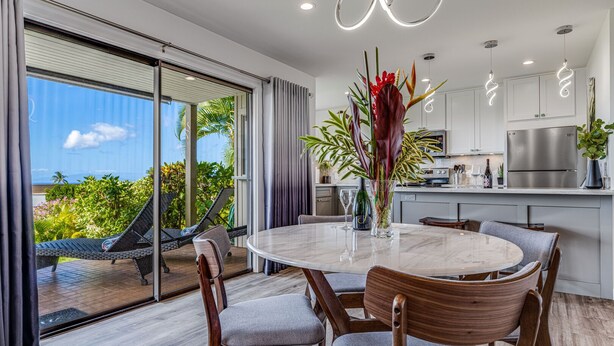 A modern kitchen with a stainless steel refrigerator, a dining area with a round table and chairs, and a view of the outdoors through large sliding doors. - Wailea Ekolu – Oceanview in the Heart of Wailea (Kihei)