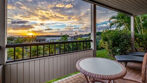 A balcony with a view of a sunset over a cityscape, surrounded by greenery and furnished with wicker furniture. - Wailea Ekolu – Oceanview in the Heart of Wailea (Kihei)