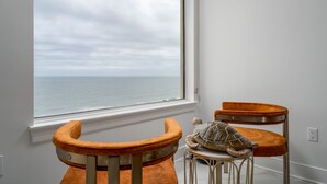Una habitación con vista al mar a través de un ventanal amplio, una silla de madera y una mesita con un cojín.