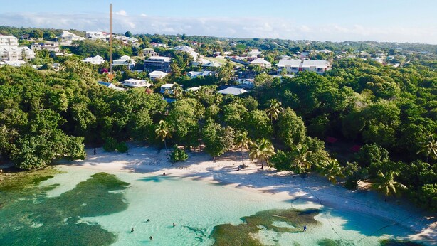 Luchtbeeld van een kustgebied met helder turquoise water, weelderige begroeiing en diverse gebouwen, waaronder woonhuizen en mogelijk ook bedrijfsgebouwen.