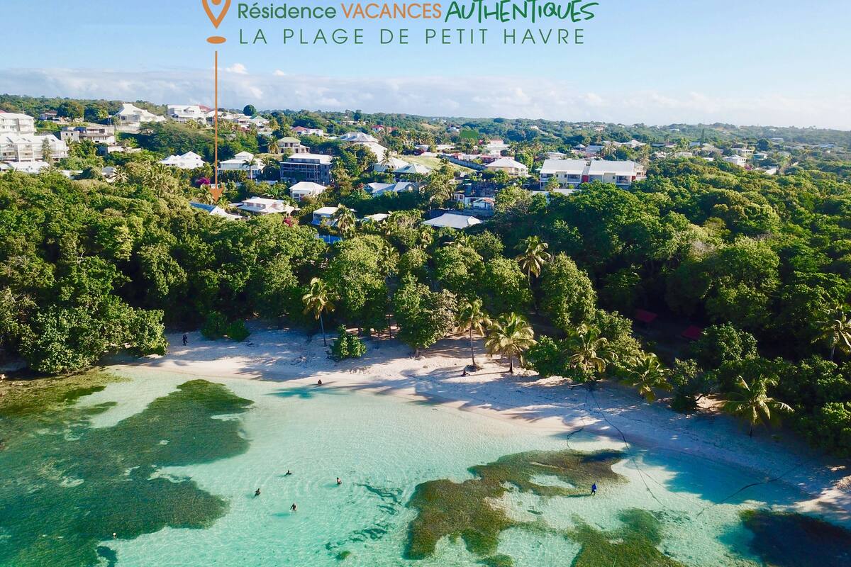 Vue aérienne d’une zone côtière caractérisée par des eaux turquoise limpides, une végétation luxuriante et une variété de bâtiments, notamment des habitations et, possiblement, des structures commerciales.