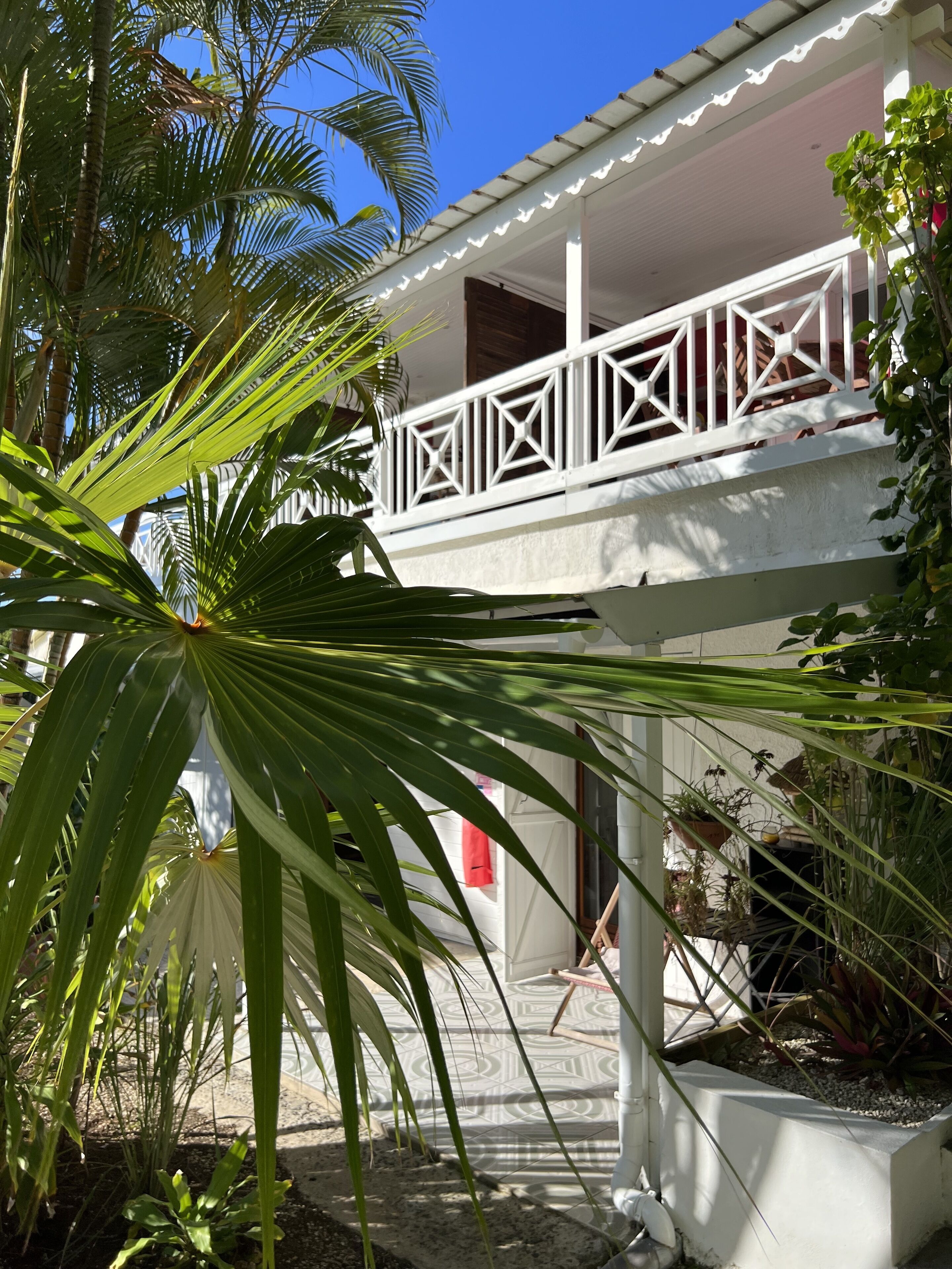 Un bâtiment à deux étages, à l’extérieur blanc, doté d’un balcon avec une balustrade aux motifs géométriques et d’un jardin agrémenté de diverses plantes.