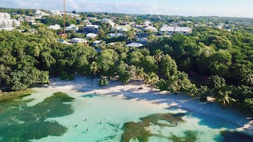 An aerial view of a coastal area with clear turquoise waters, dense greenery, and a variety of buildings, including residential and possibly commercial structures.