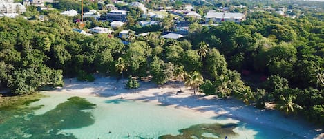 Vista aérea de una zona costera con aguas turquesas cristalinas, abundante vegetación y diversos edificios, incluyendo residencias y posiblemente establecimientos comerciales.
