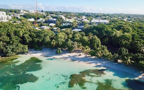 Luchtbeeld van een kustgebied met helder turquoise water, weelderige begroeiing en diverse gebouwen, waaronder woonhuizen en mogelijk ook bedrijfsgebouwen.