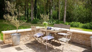 Une terrasse extérieure avec une table en pierre et des chaises en métal, entourée d’un mur de pierre et d’une zone boisée.