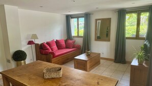 Una habitación con una mesa de madera, un sofá rojo, una mesita auxiliar de madera, una lámpara, un espejo y una ventana con cortinas.