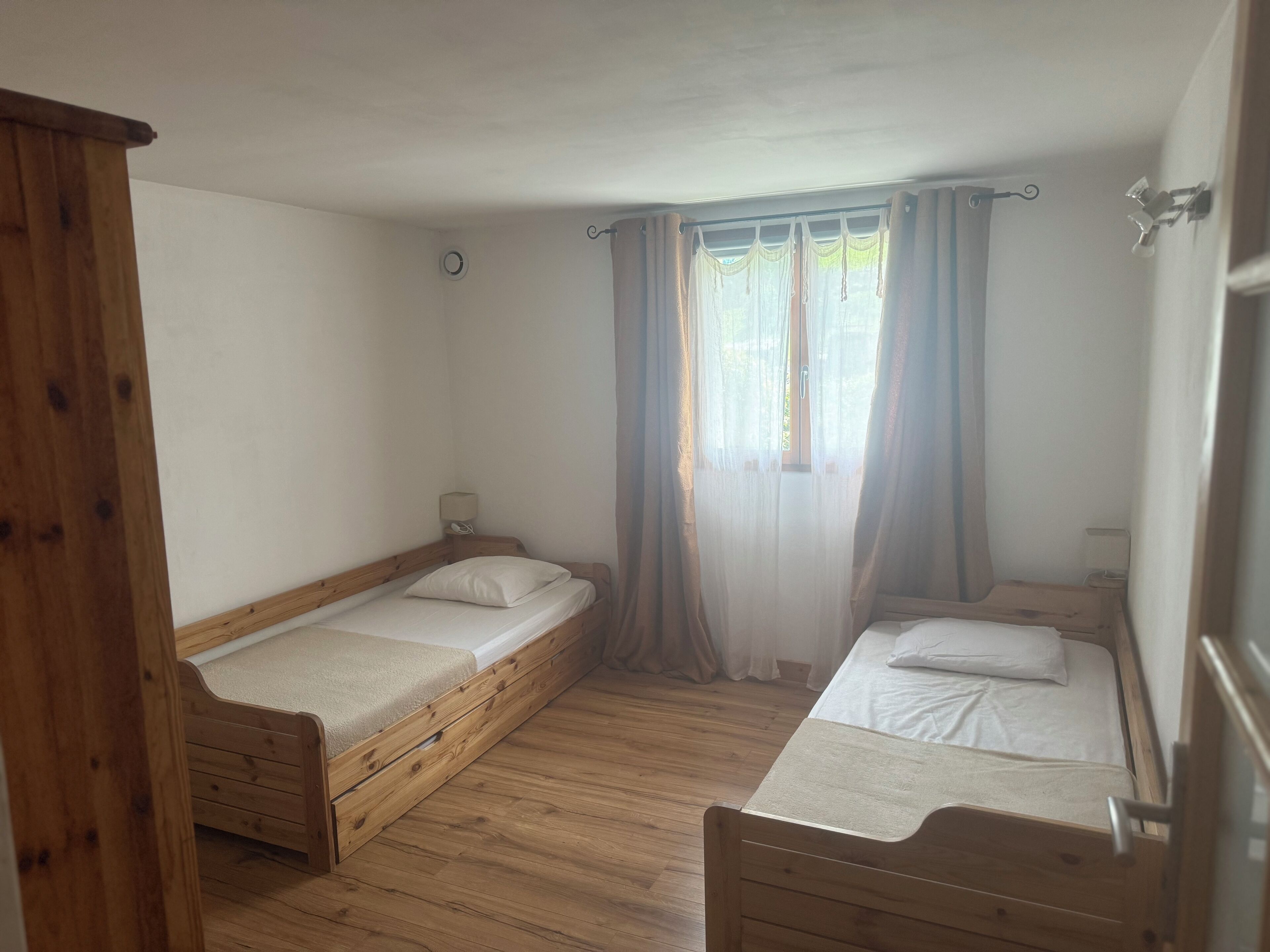 Ein Zimmer mit zwei Holzbettgestellen, einem Schrank aus Holz, einem kleinen Nachttisch mit Lampe und einem Fenster mit transparenten Vorhängen.