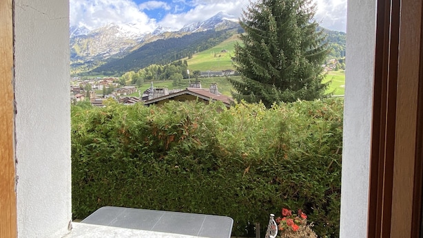 Vista desde una ventana sobre un paisaje montañoso con un pueblo abajo, un gran árbol de hoja perenne y una silla metálica en primer plano.