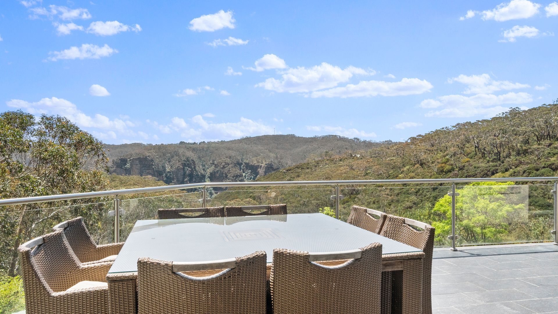 A dining area with a glass-top table and wicker chairs, overlooking a mountainous landscape.