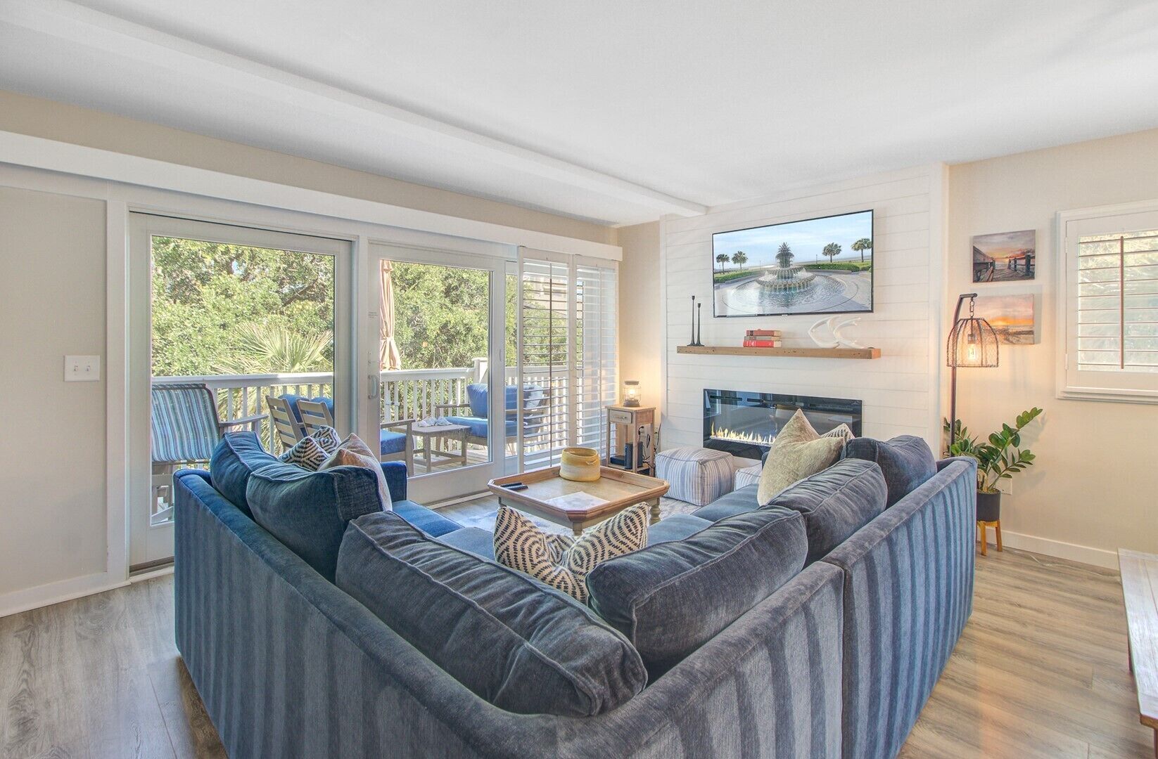 A living room with a large sectional sofa, a fireplace, a television mounted on the wall, and a balcony with outdoor seating.