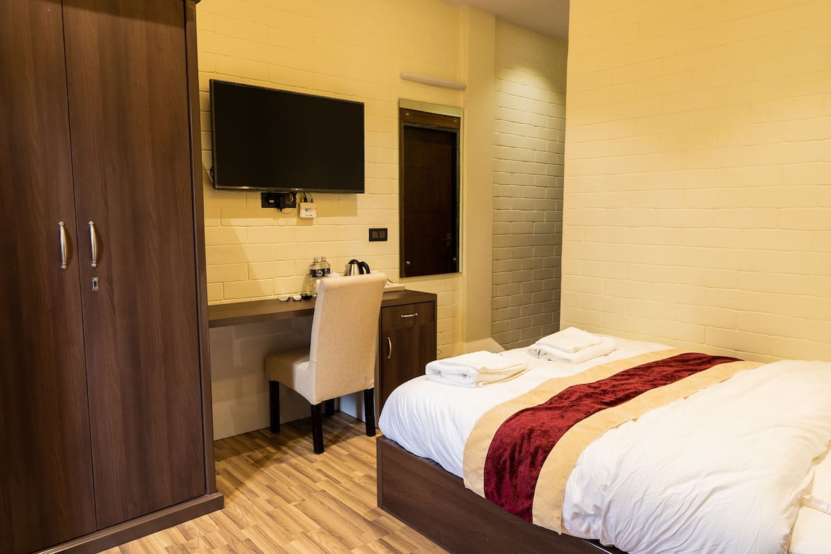 Habitación de hotel con armario de madera, escritorio y silla, cama con ropa blanca y una manta roja, y televisor de pantalla plana montado en la pared.