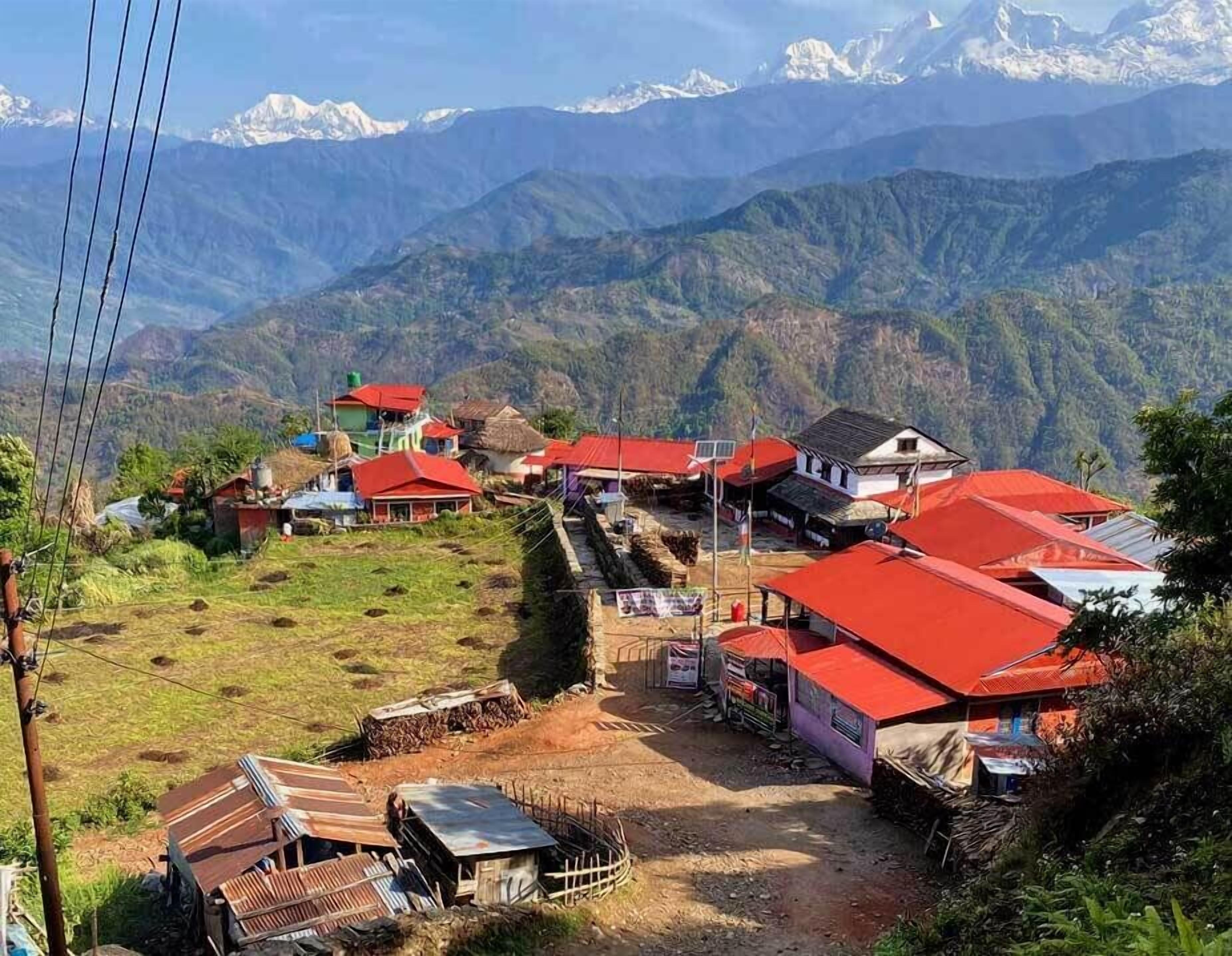A mountainous village with houses having red roofs, a dirt road, and a clear view of snow-capped mountains in the background.