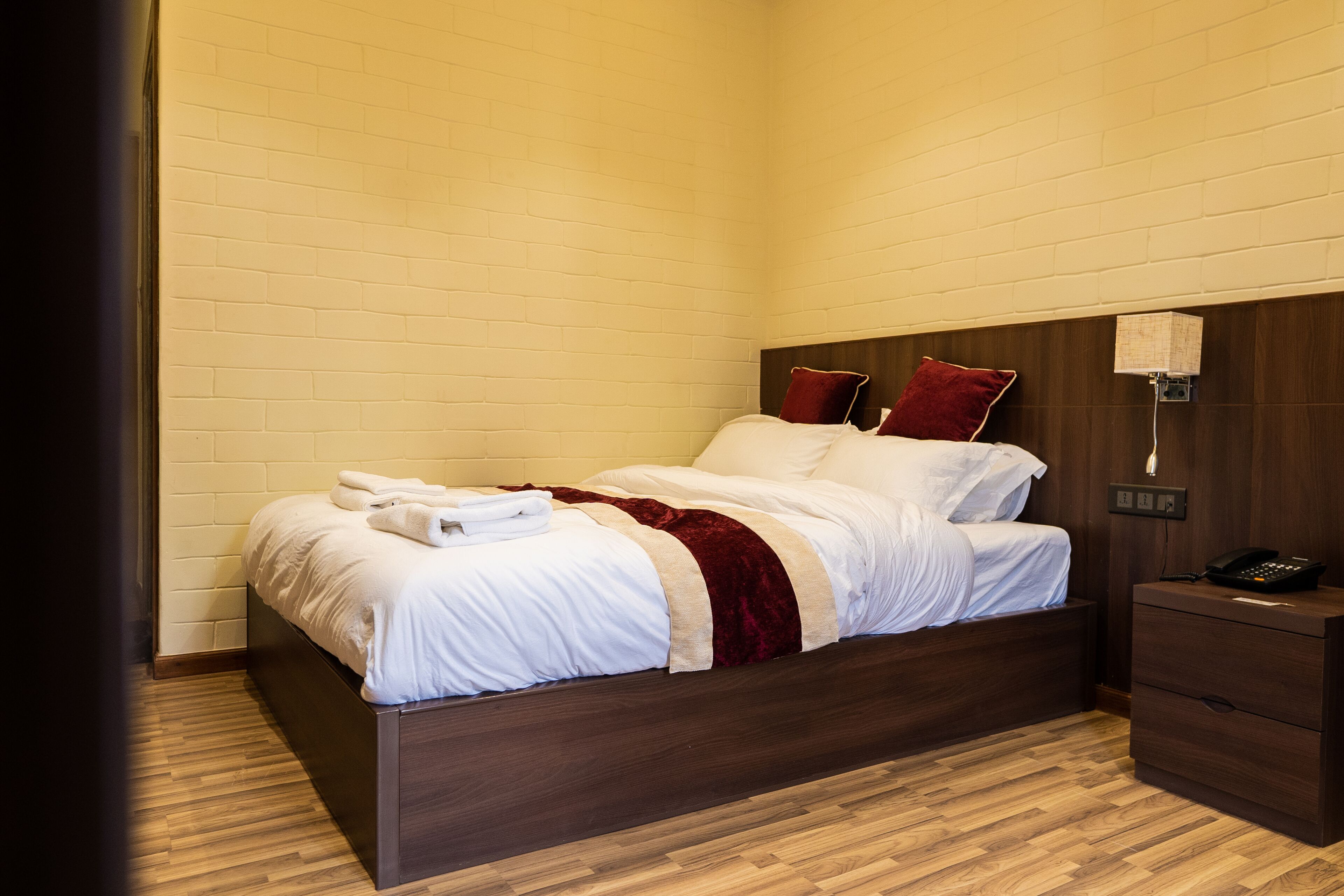 Un lit bien fait, avec du linge de lit blanc et un plaid rouge, une table de chevet en bois avec un téléphone, et un luminaire fixé au mur.