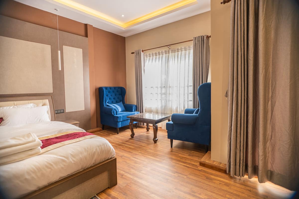 Habitación de hotel con una cama grande, dos sillones azules, una mesita de madera y un ventanal con cortinas.