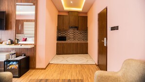 Una habitación de hotel compacta con kitchenette, televisor de pantalla plana, cama con almohadas, una pequeña zona de estar y suelo de madera.
