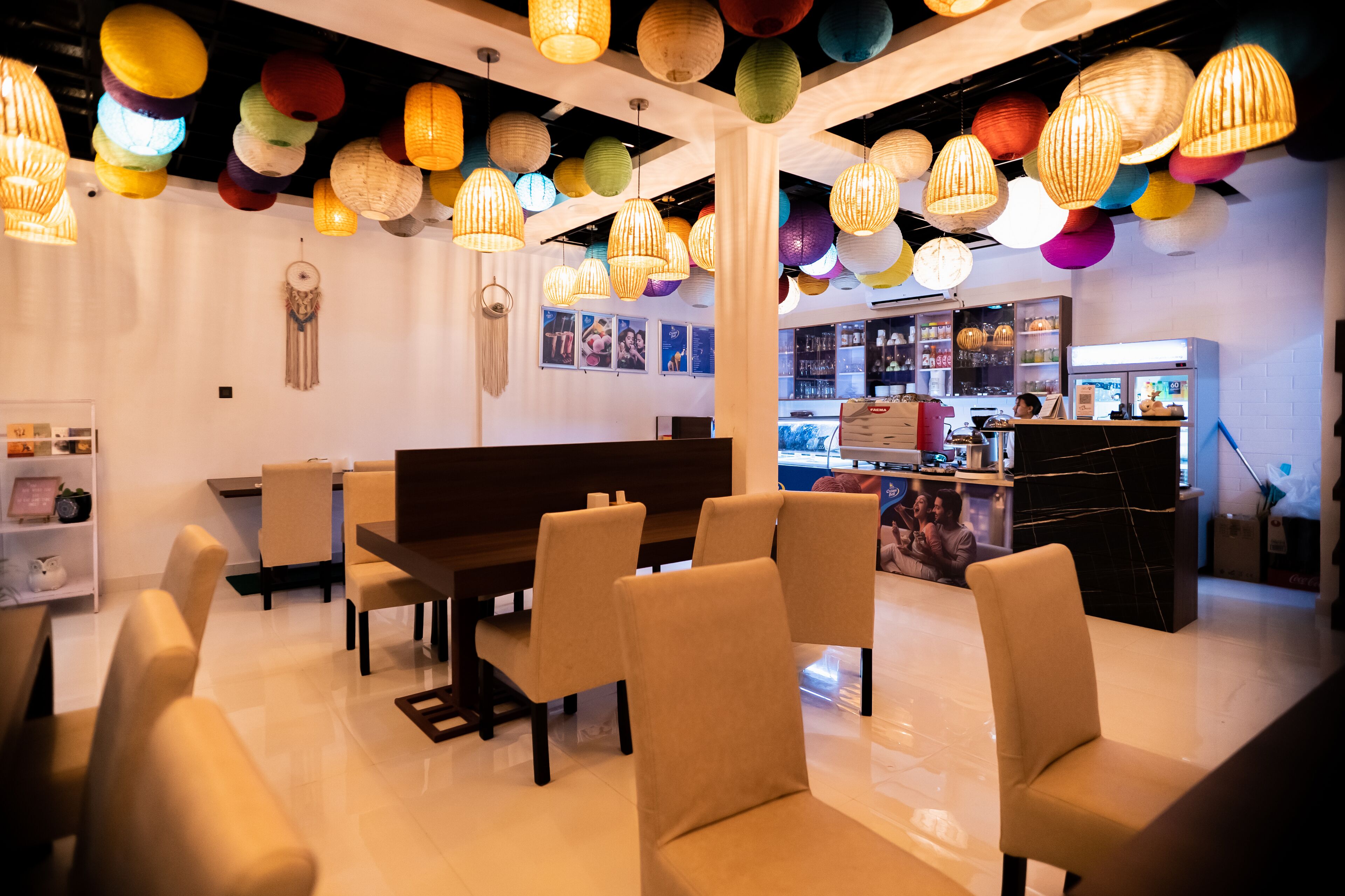 A modern interior with a dining area featuring beige chairs and a wooden table, a bar area with a counter and seating, and colorful hanging paper lanterns.