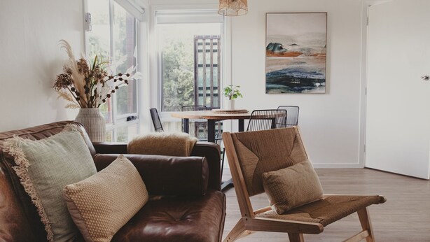 Een moderne woonkamer met een bruine leren bank, een geweven stoel en een eettafel met stoelen. Er is een groot raam met uitzicht op een gebouw.