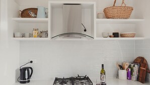 Een keuken met witte kastjes, een gasfornuis, een waterkoker en diverse keukenspullen op planken.