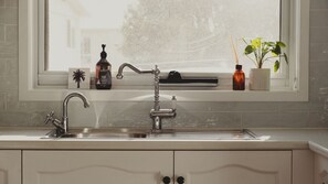 A kitchen with a double sink, a window above the sink, and a shelf with various items including a bottle, a plant, and a small figurine. - Reardon Retreat - A stones throw to the beach (Port Fairy)