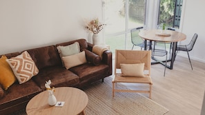 A modern living room with a brown leather sofa, a wooden chair, a small round table, and a large window offering a view of the outdoors. - Reardon Retreat - A stones throw to the beach (Port Fairy)