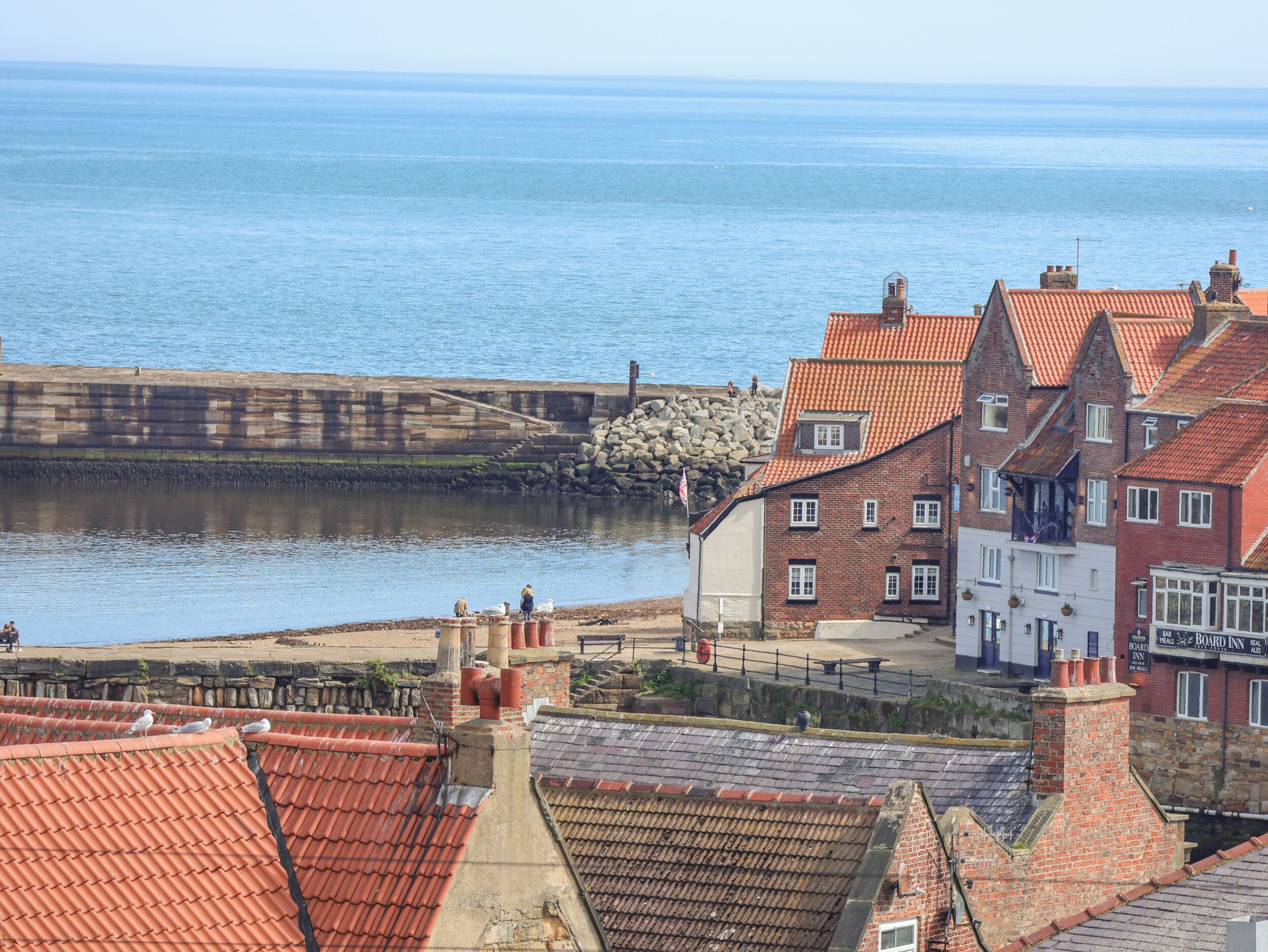 A coastal town with traditional red-tiled buildings, a stone pier, and a calm sea.