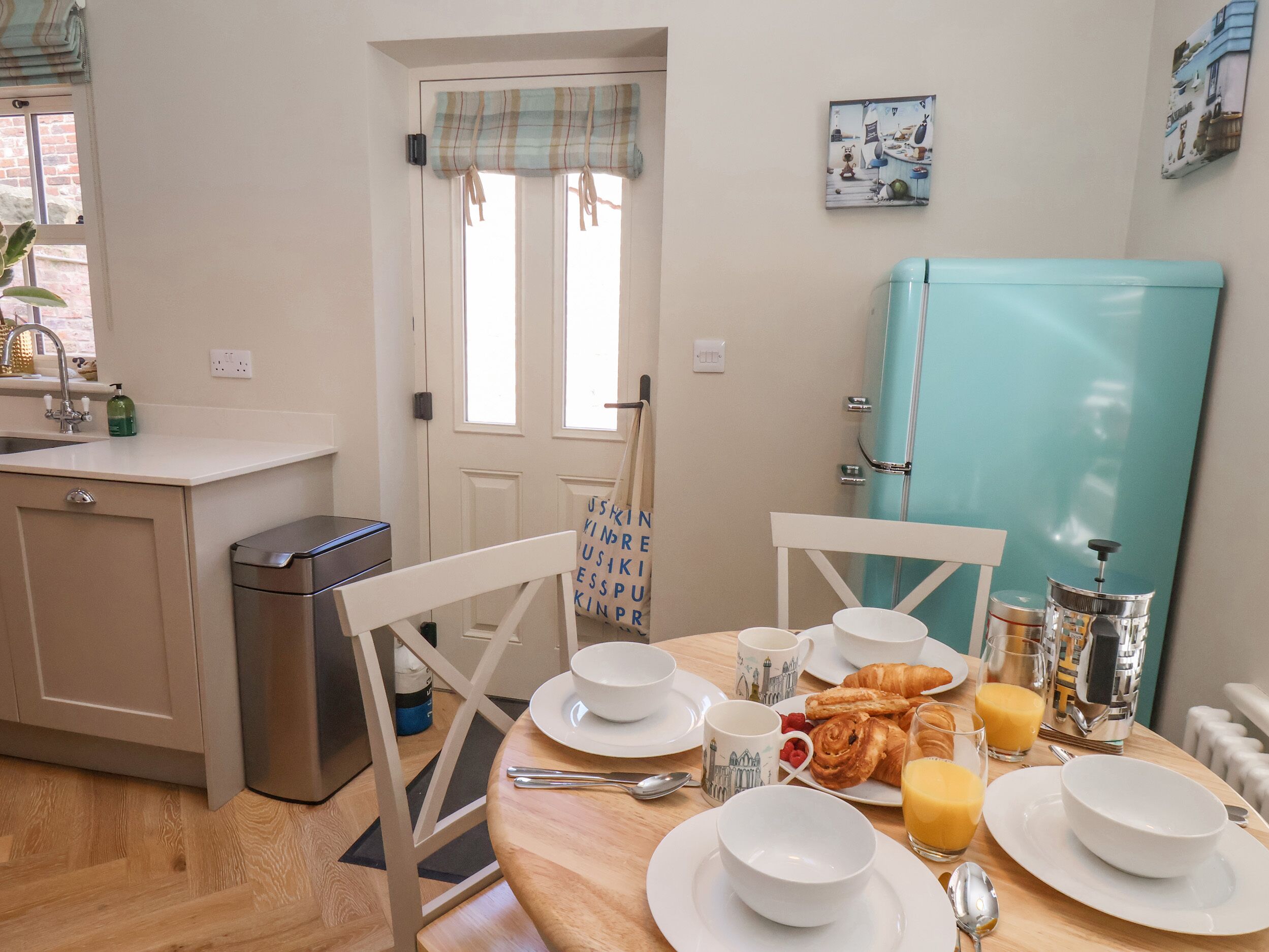 A breakfast setup with plates, cups, croissants, and juice in a room with a kitchen counter, a sink, and a teal refrigerator.