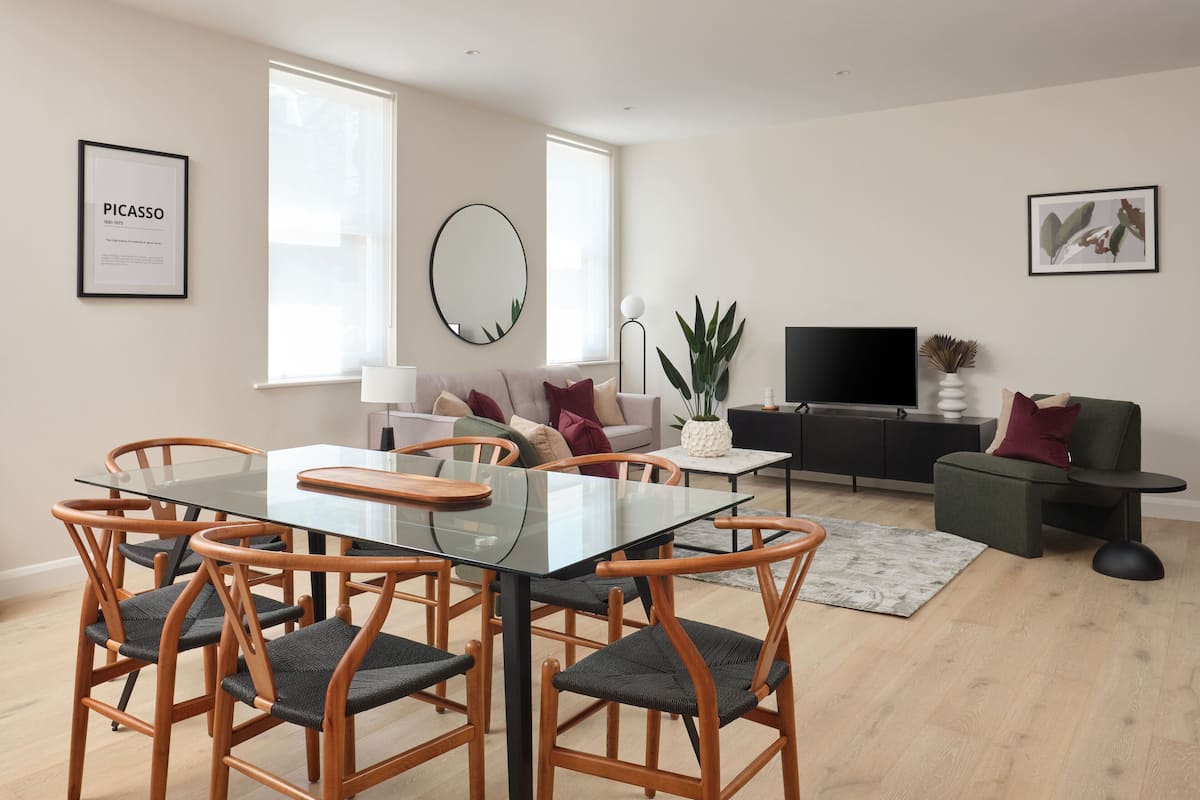 Un salon moderne avec une table à manger au plateau en verre, des chaises en bois, un canapé, un téléviseur et des œuvres d’art accrochées aux murs.