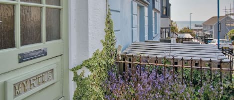 A quaint townhouse with a green door labeled "MYRTLE," a wooden fence, and a garden with purple flowers.