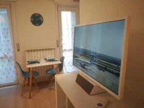 Una habitación con una televisión de pantalla plana grande sobre un mueble, una mesa de comedor con sillas, y un reloj de pared.