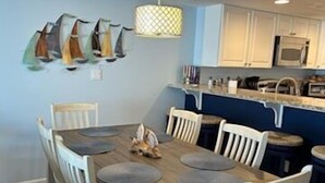 A dining area with a wooden table and white chairs, a hanging light fixture, and a kitchen in the background.
