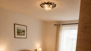Een houten bed met twee kussens, een nachtkastje met een lamp, een ingelijst foto aan de muur en een raam met gordijnen.
