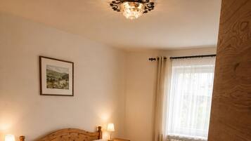 A wooden bed with two pillows, a bedside table with a lamp, a framed picture on the wall, and a window with curtains.