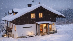 Een houten hutje met een met sneeuw bedekte dak en een verlichte ingang, gelegen in een besneeerd landschap.