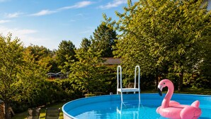 Een bovengronds zwembad met een ladder, een roze flamingo drijvend in het water en ligstoelen op een gazon.
