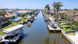 A canal with boats docked on both sides, surrounded by residential buildings and palm trees. - Feeling TipSea | Heated Pool | Gulf Access & Dock (Cape Coral)