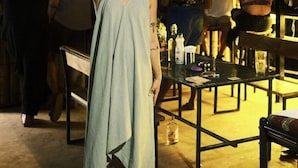 A person in a light blue dress standing in an outdoor dining area with tables and chairs, a thatched roof, and a chalkboard menu in the background. - SPACELAND Mandrem (Mandrem)