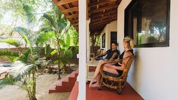 A covered outdoor seating area with wicker chairs, a white wall, and a window overlooking a garden with palm trees.