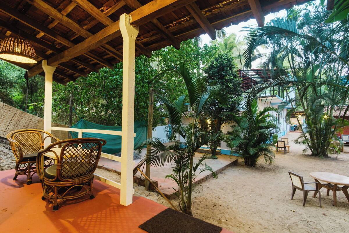 Un espacio al aire libre con sillas de mimbre y una mesa, rodeado de palmeras y una pérgola de madera.