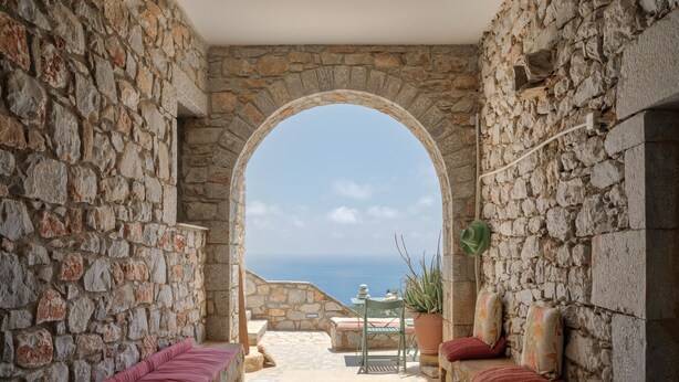 Un couloir aux murs de pierre donnant sur la mer, une table avec des chaises et une plante en pot.