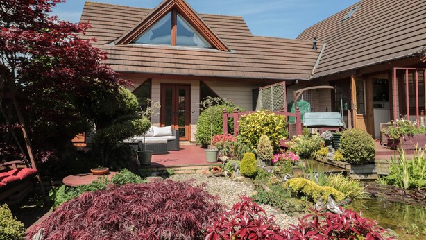 A house with a sloped roof, a front porch, and a garden with various plants and a pond. - Swallow Lodge (Morpeth)