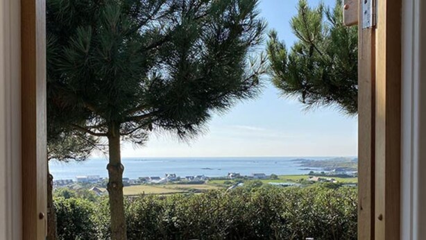 A wooden deck with a table and chairs, overlooking a coastal landscape with trees and a view of the sea.