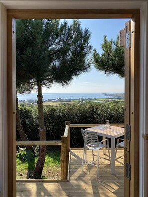 Un pontile in legno con tavolo e sedie, affacciato su un paesaggio costiero con alberi e vista sul mare.