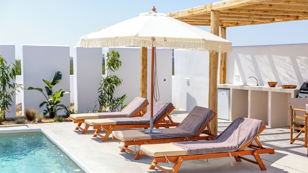 Un área junto a la piscina con sillones de madera, una sombrilla blanca y una zona de bar.