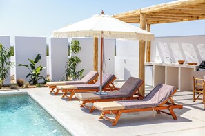 Un espace aménagé au bord de la piscine, avec des chaises longues en bois, un parasol blanc et un coin bar.