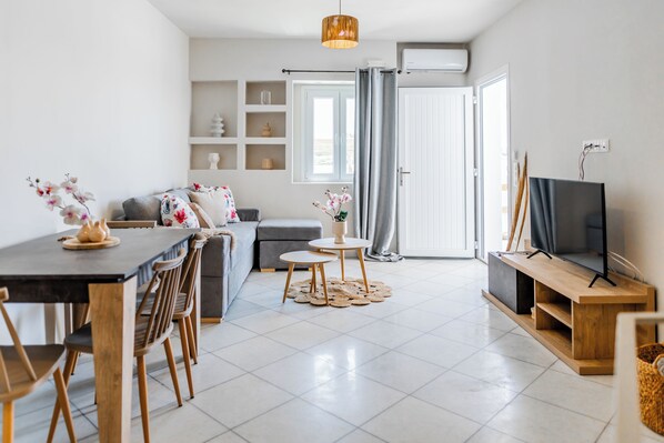 Un salon moderne comprenant un coin repas, un canapé gris, un téléviseur et une grande fenêtre avec des rideaux.