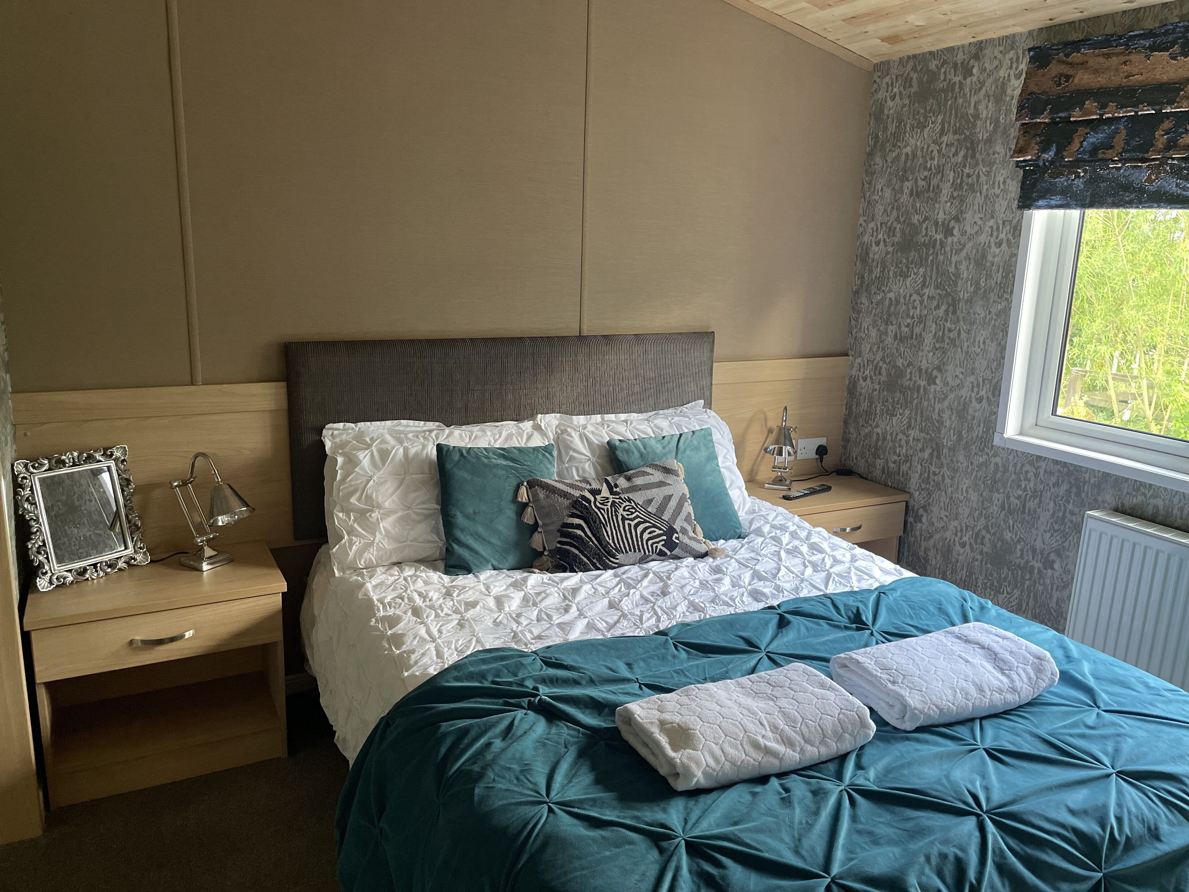 A bedroom with a bed, a nightstand, a lamp, a picture frame, and a window with a view of greenery.