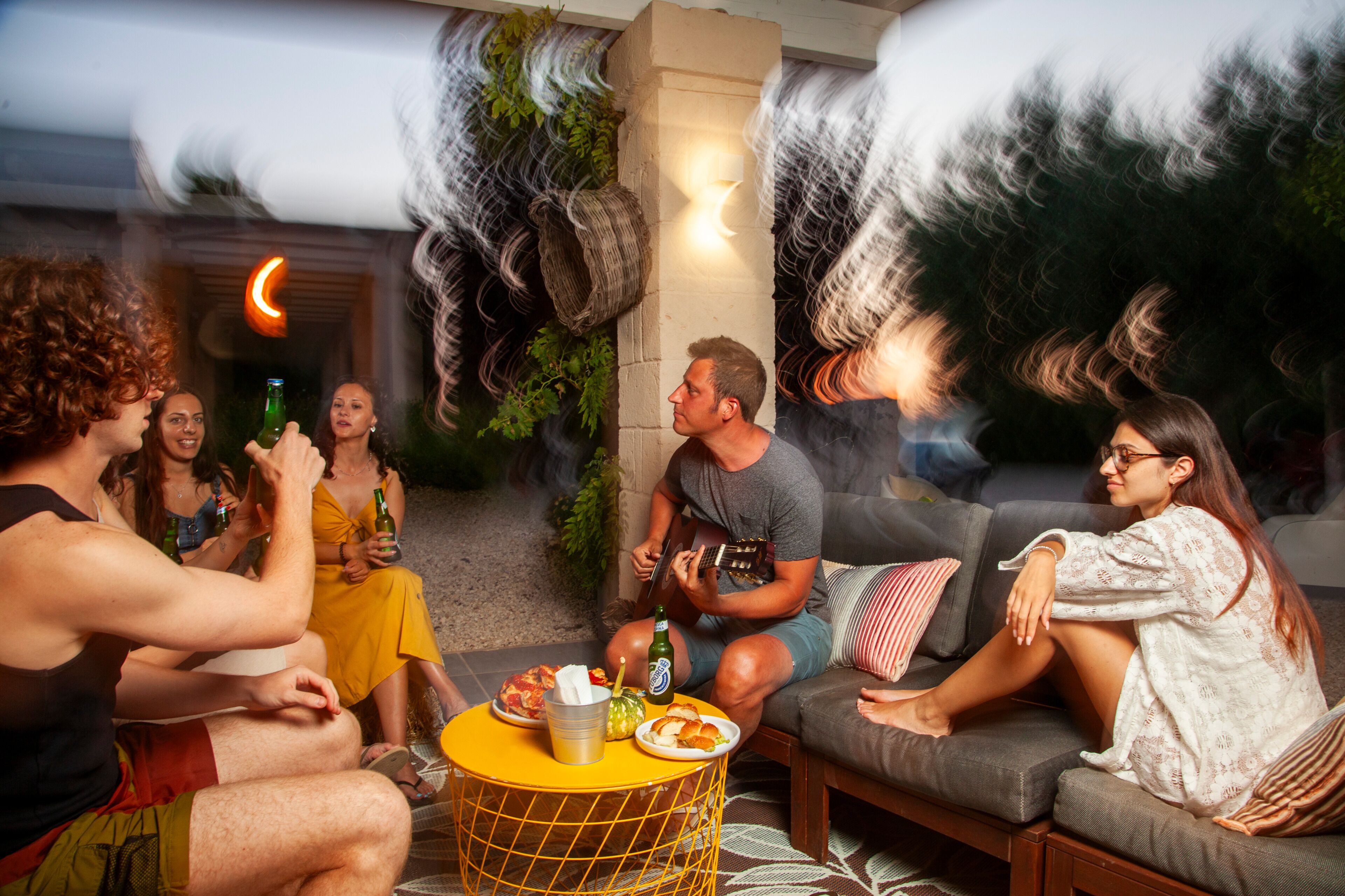 A group of people sitting on a couch and chairs in a living room with a yellow coffee table, a bottle of beer, and a plate of food.