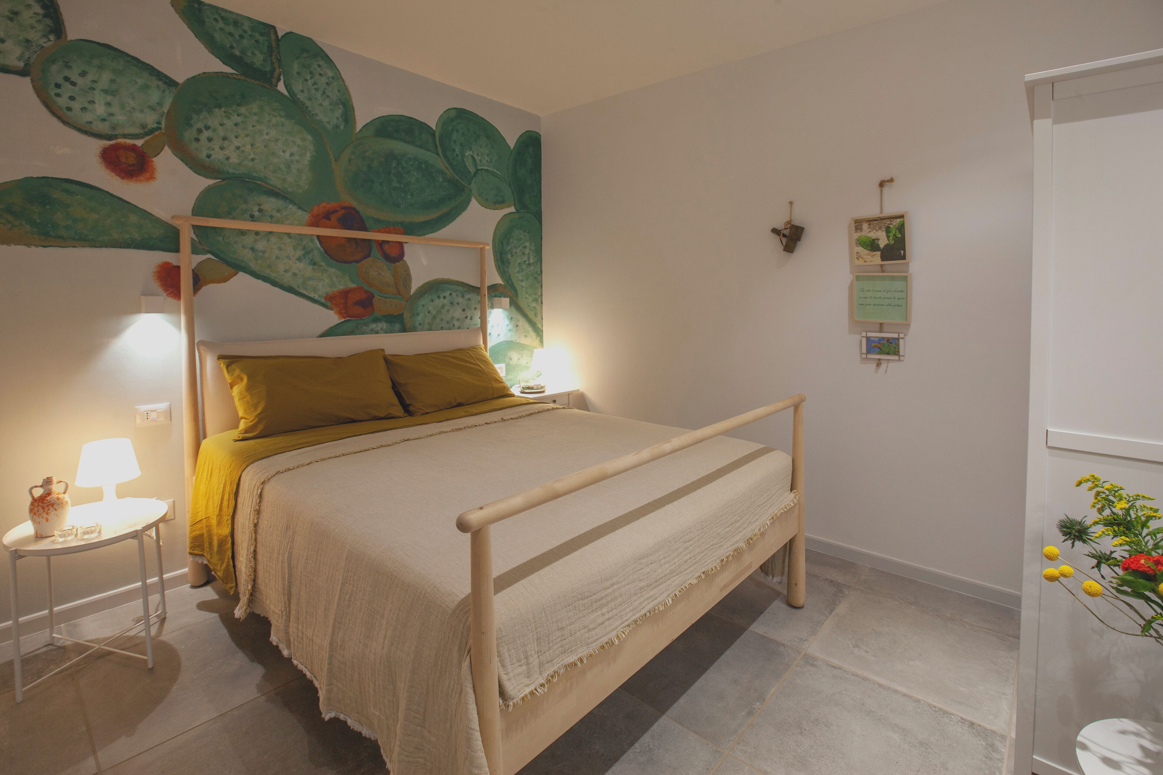 A bed with a wooden frame, a yellow pillow, and a white bedspread. There is a small table with a lamp and a vase with flowers.