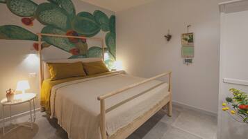 A bed with a wooden frame, a yellow pillow, and a white bedspread. There is a small table with a lamp and a vase with flowers.